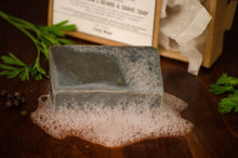 Grey shaving soap bar with foam beneath and green leaves and box in the background