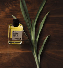 Rectangular clear beard oil bottle filled with yellow beard oil alongside green plant leaves