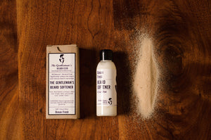 Bottle of beard softener laying flat next to a box and a pile of beach sand