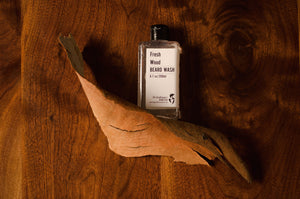 Beard wash bottle laying flat on a wooden surface next to a piece of tree bark