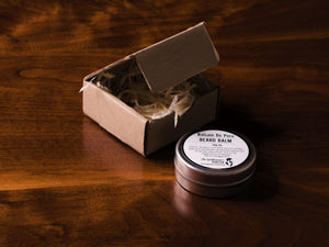 Tin of beard balm laying flat on a wooden surface with an open brown packaging box in the background